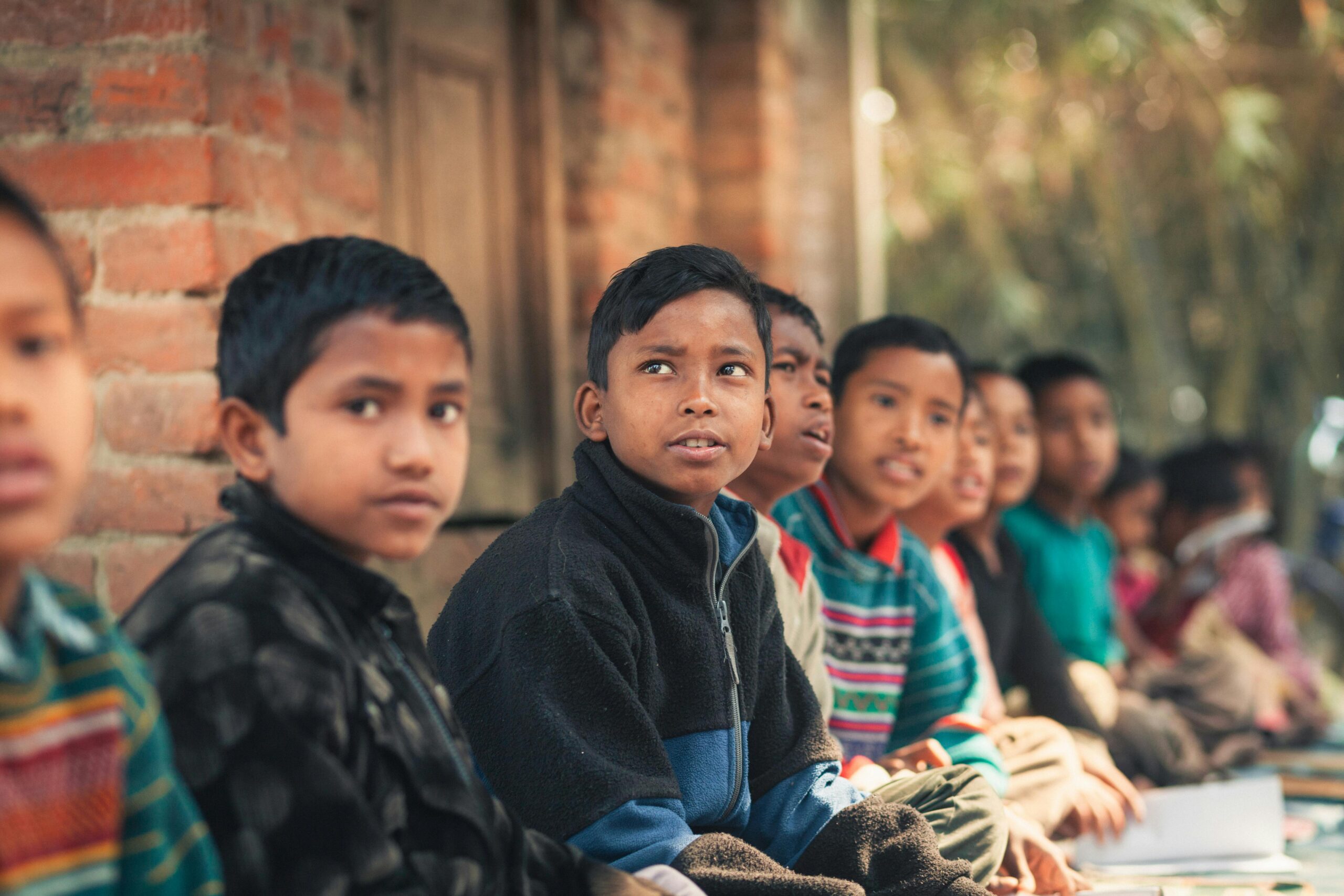 Home A group of children from Bangladesh sitting outdoors, engaged in learning at a rural school.